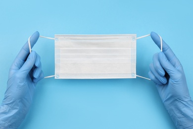 Doctor in medical gloves holding protective mask on light blue background, closeup