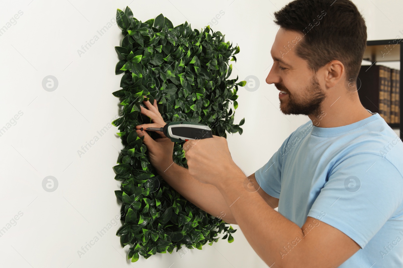 Photo of Man with screwdriver installing green artificial plant panel on white wall in room