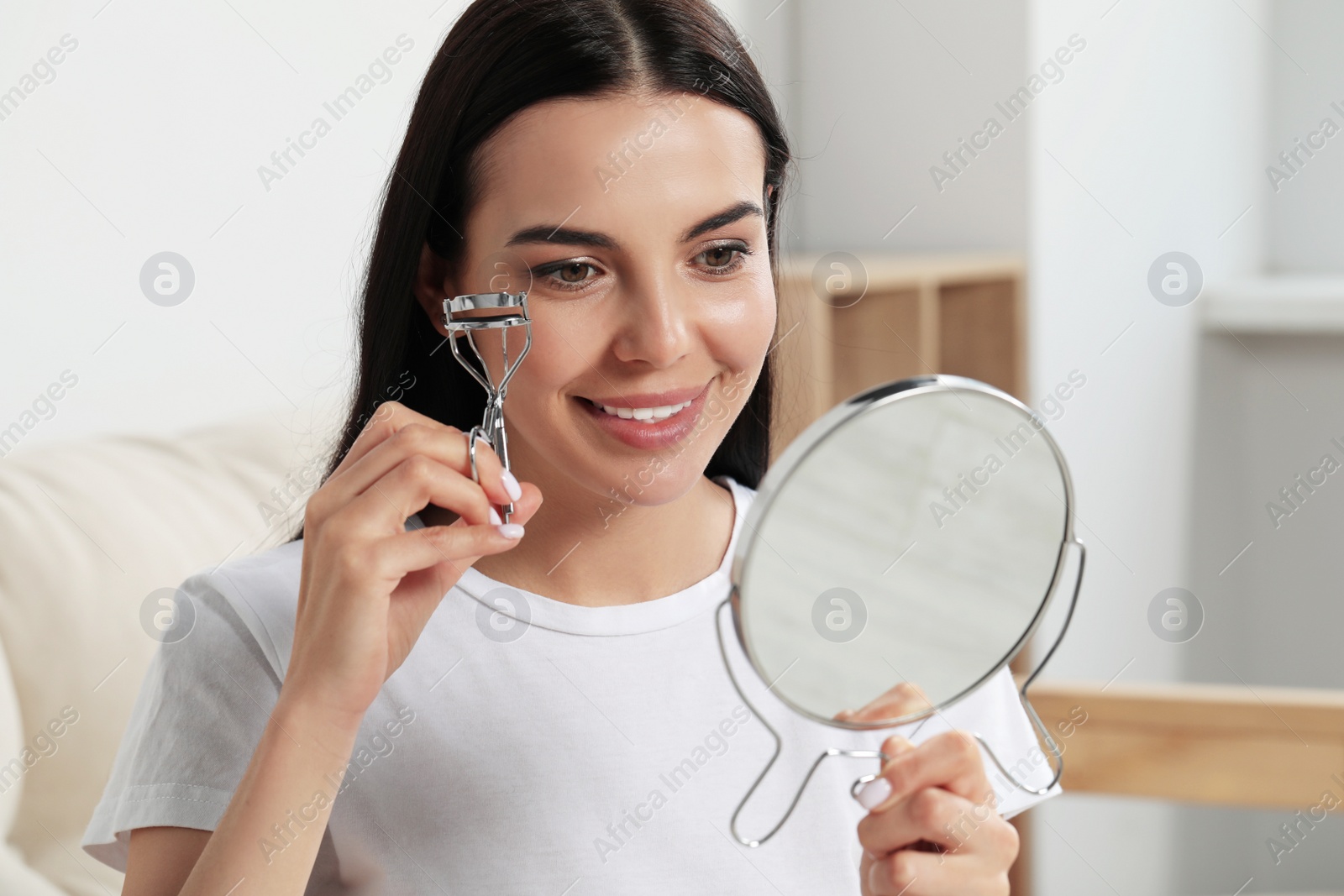 Photo of Beautiful young woman with mirror using eyelash curler indoors