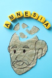 Photo of Yellow cubes with word Amnesia and human head with brain pieces made of crumbled paper on light blue background, flat lay