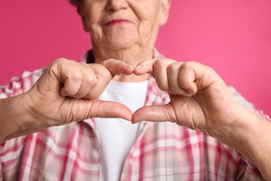 Elderly woman making heart with her hands on pink background, closeup