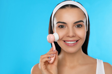 Photo of Young woman using facial cleansing brush on light blue background, space for text. Washing accessory