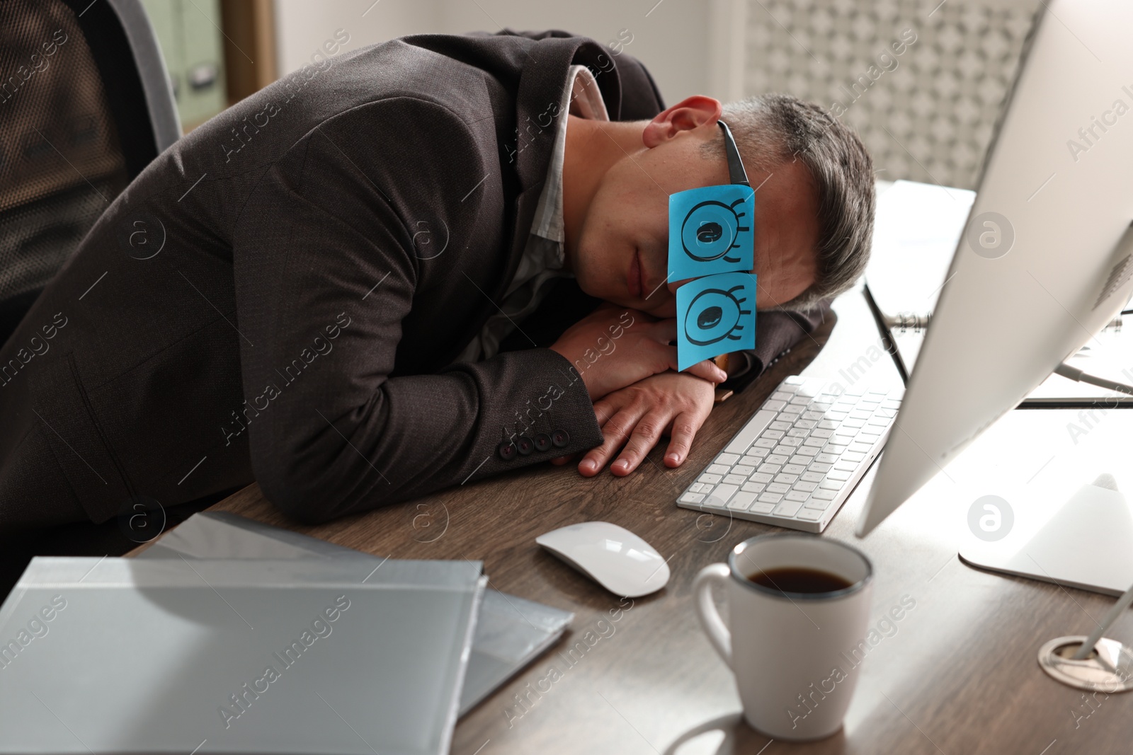 Photo of Man with fake eyes painted on sticky notes snoozing at workplace in office