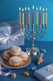 Hanukkah celebration. Menorah with burning candles, dreidels, donuts and gift box on light blue table