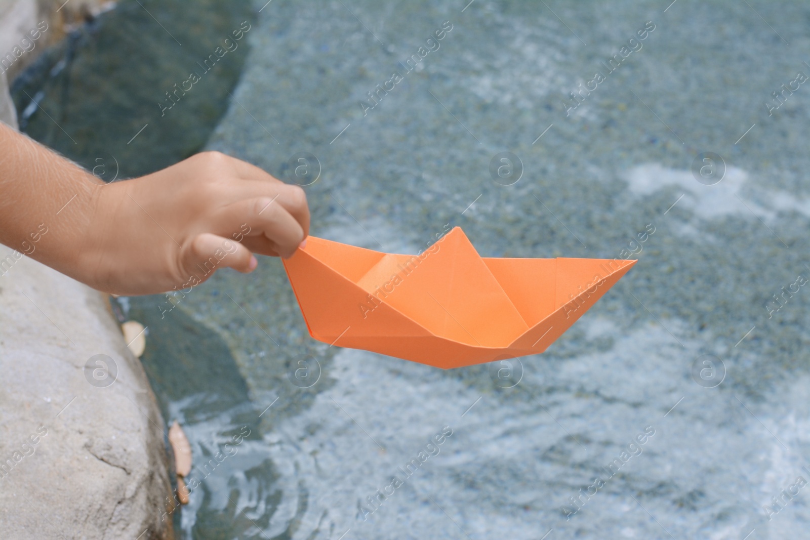 Photo of Kid launching small orange paper boat on water outdoors, closeup