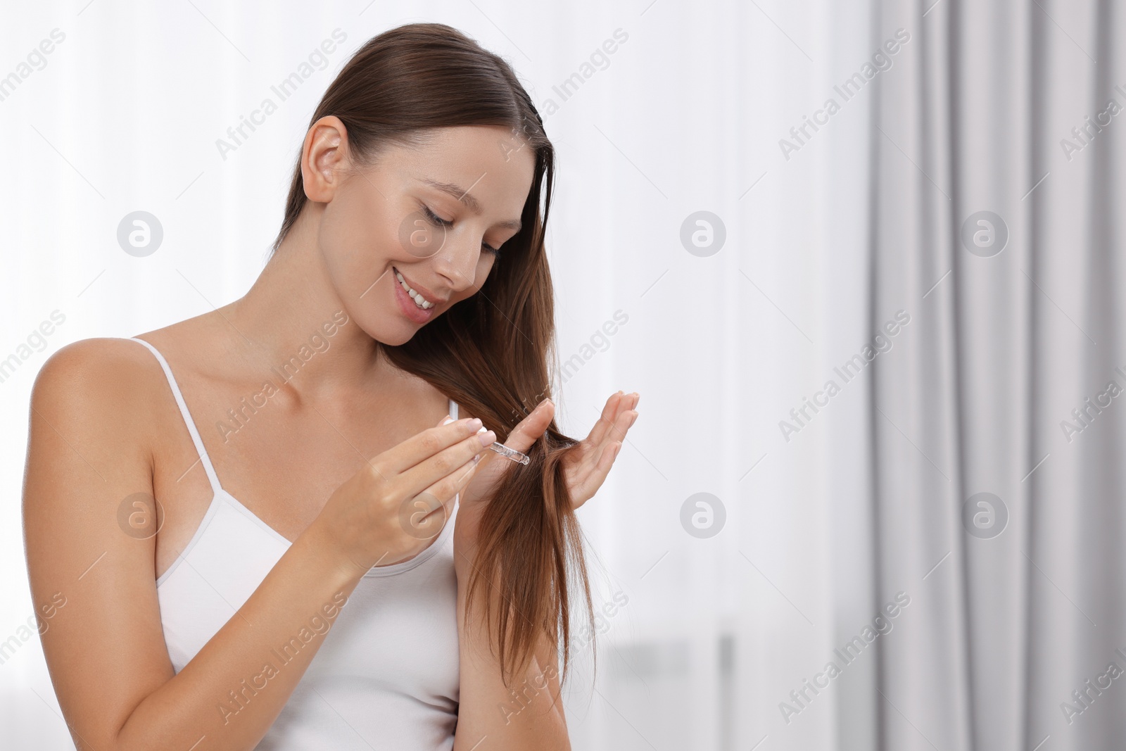 Photo of Beautiful woman applying serum onto hair indoors, space for text