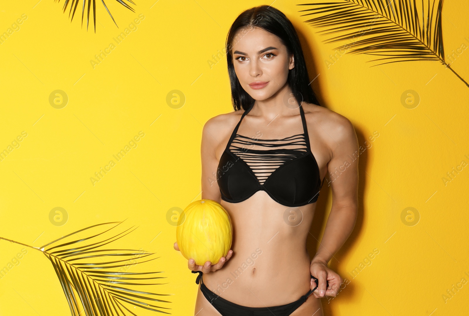 Photo of Beautiful young woman in black bikini with melon and tropical leaves on yellow background