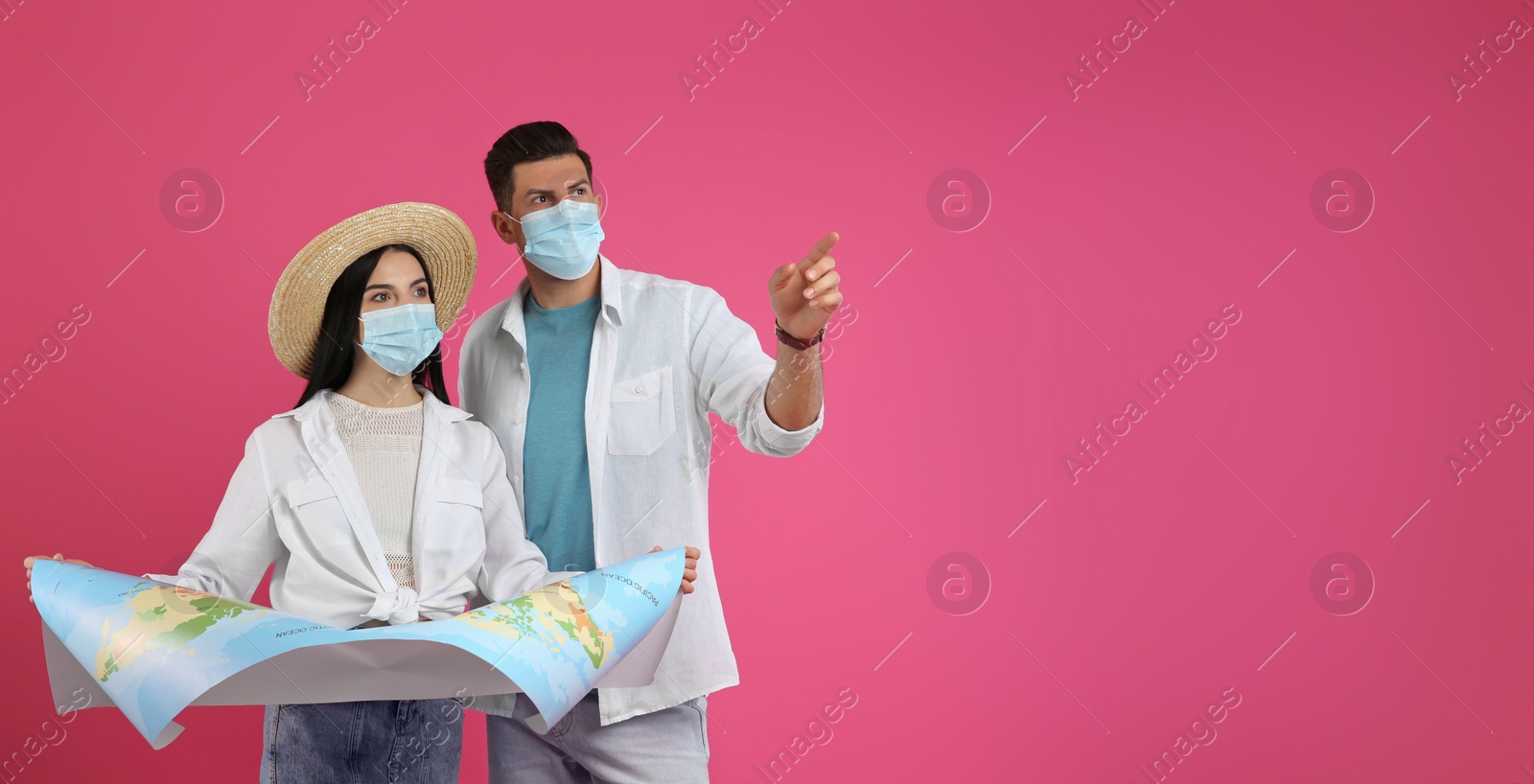 Photo of Couple of tourists in medical masks with map on pink background, space for text. Travelling during coronavirus pandemic