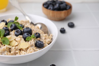 Photo of Tasty oatmeal with blueberries, mint and almond flakes in bowl on white tiled table, closeup. Space for text