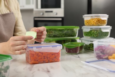 Photo of Woman sticking paper note onto container with fresh carrots at white marble table in kitchen, closeup. Food storage