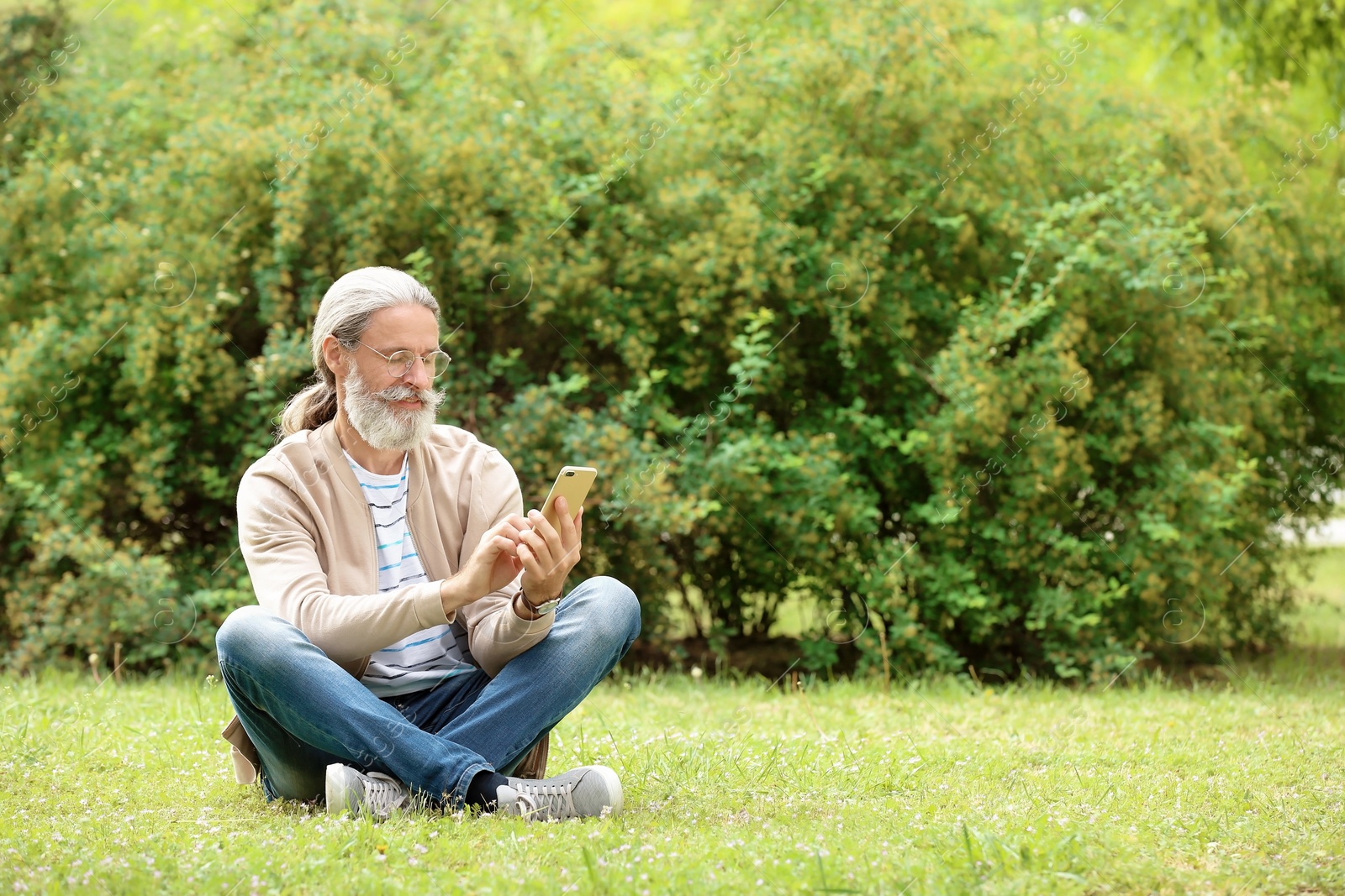 Photo of Handsome mature man with mobile phone sitting on green grass in park