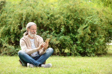 Photo of Handsome mature man with mobile phone sitting on green grass in park
