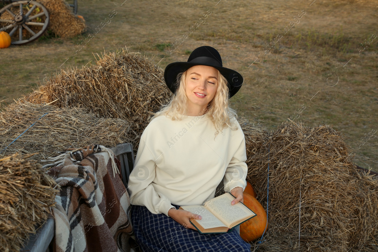 Photo of Beautiful woman with book sitting on wooden bench near hay bales outdoors. Autumn season