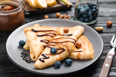 Photo of Delicious thin pancakes with chocolate, blueberries and nuts on black wooden table