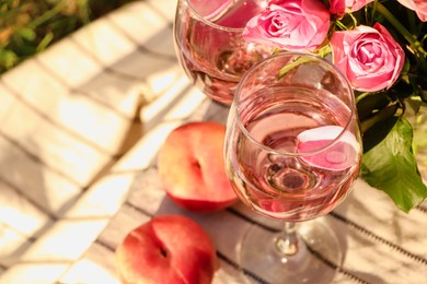 Photo of Glasses of delicious rose wine with petals, flowers and peaches on white picnic blanket outside, above view. Space for text