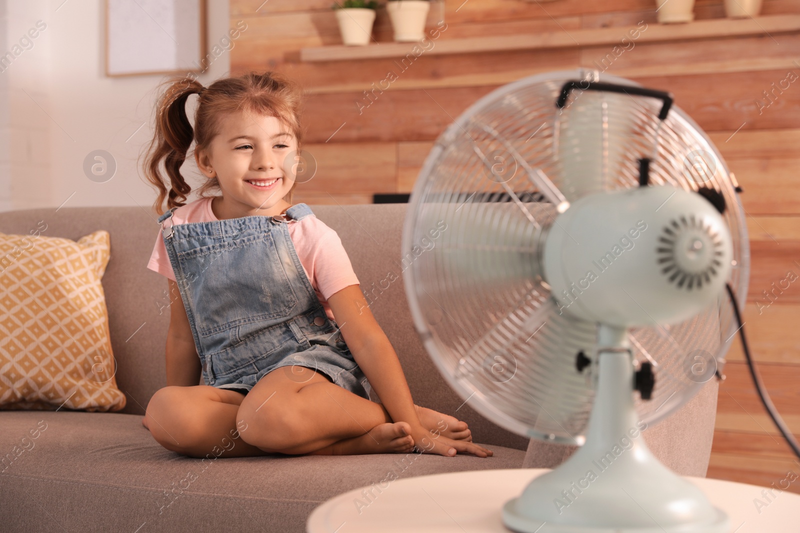 Photo of Little girl enjoying air flow from fan on sofa in living room. Summer heat