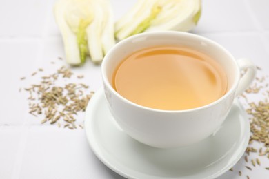 Photo of Fennel tea in cup, seeds and fresh vegetable on white tiled table, closeup