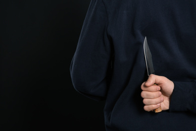 Man with knife behind his back on black background, closeup. Dangerous criminal