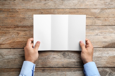 Young man holding blank brochure at wooden table, top view. Mock up for design