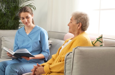 Photo of Nurse reading book to elderly woman indoors. Assisting senior people