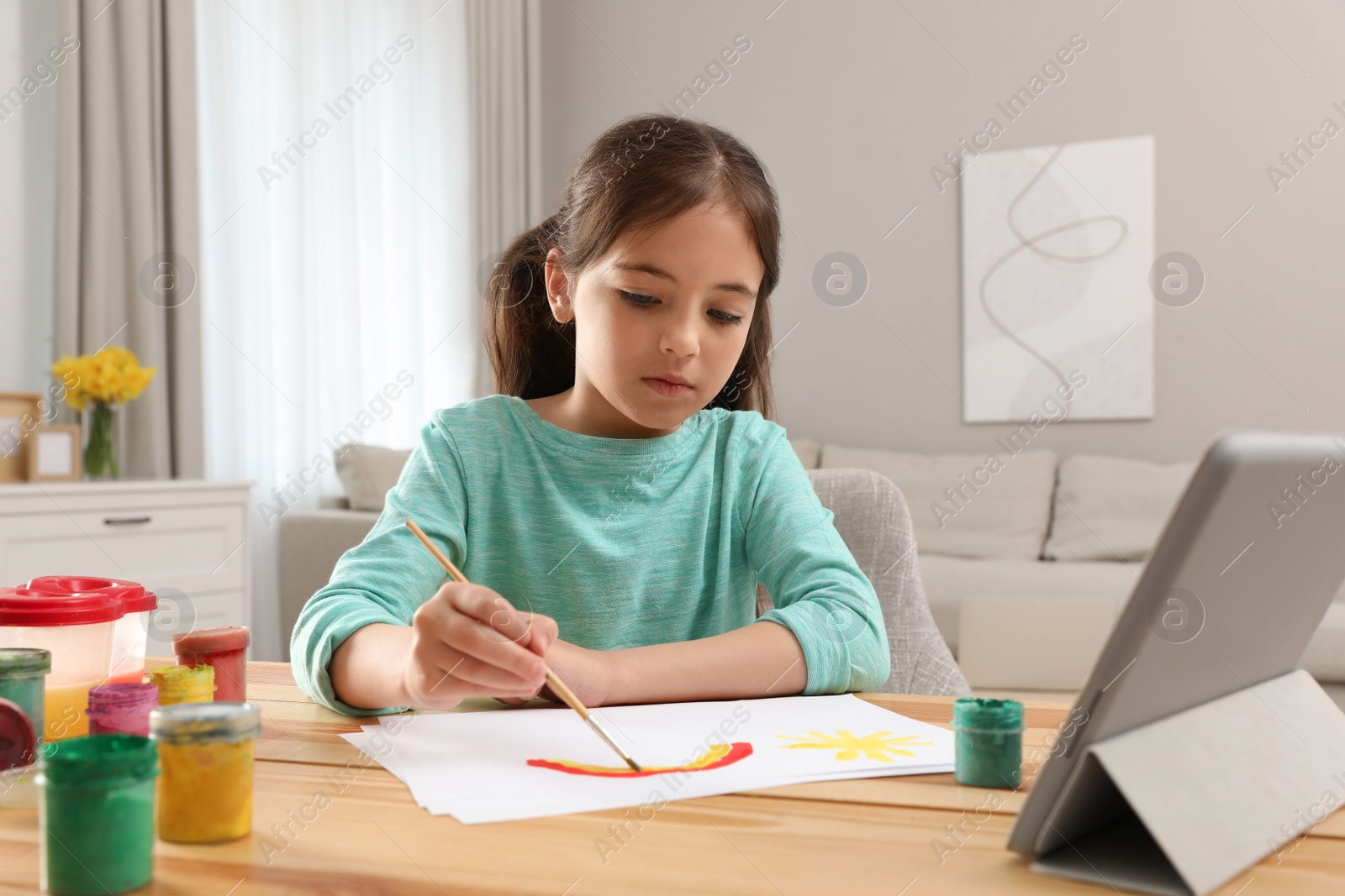 Photo of Little girl drawing on paper with paints at online lesson indoors. Distance learning