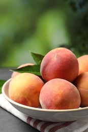 Photo of Fresh peaches and leaves in bowl on grey table against blurred green background, closeup
