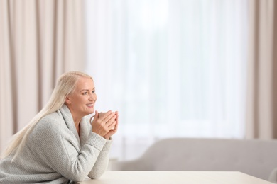 Portrait of beautiful older woman with cup of tea sitting at table indoors. Space for text