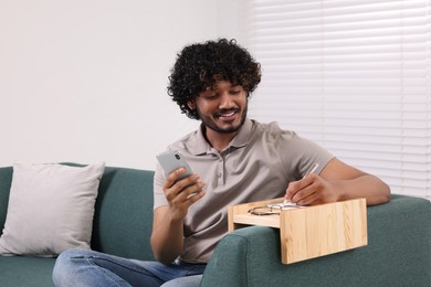 Photo of Happy man holding smartphone while writing in notebook on sofa armrest wooden table at home