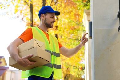 Photo of Courier in uniform with parcels ringing doorbell outdoors