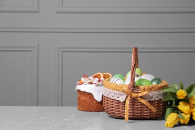 Photo of Easter basket with many painted eggs near tasty cake and bouquet of tulips on grey textured table. Space for text