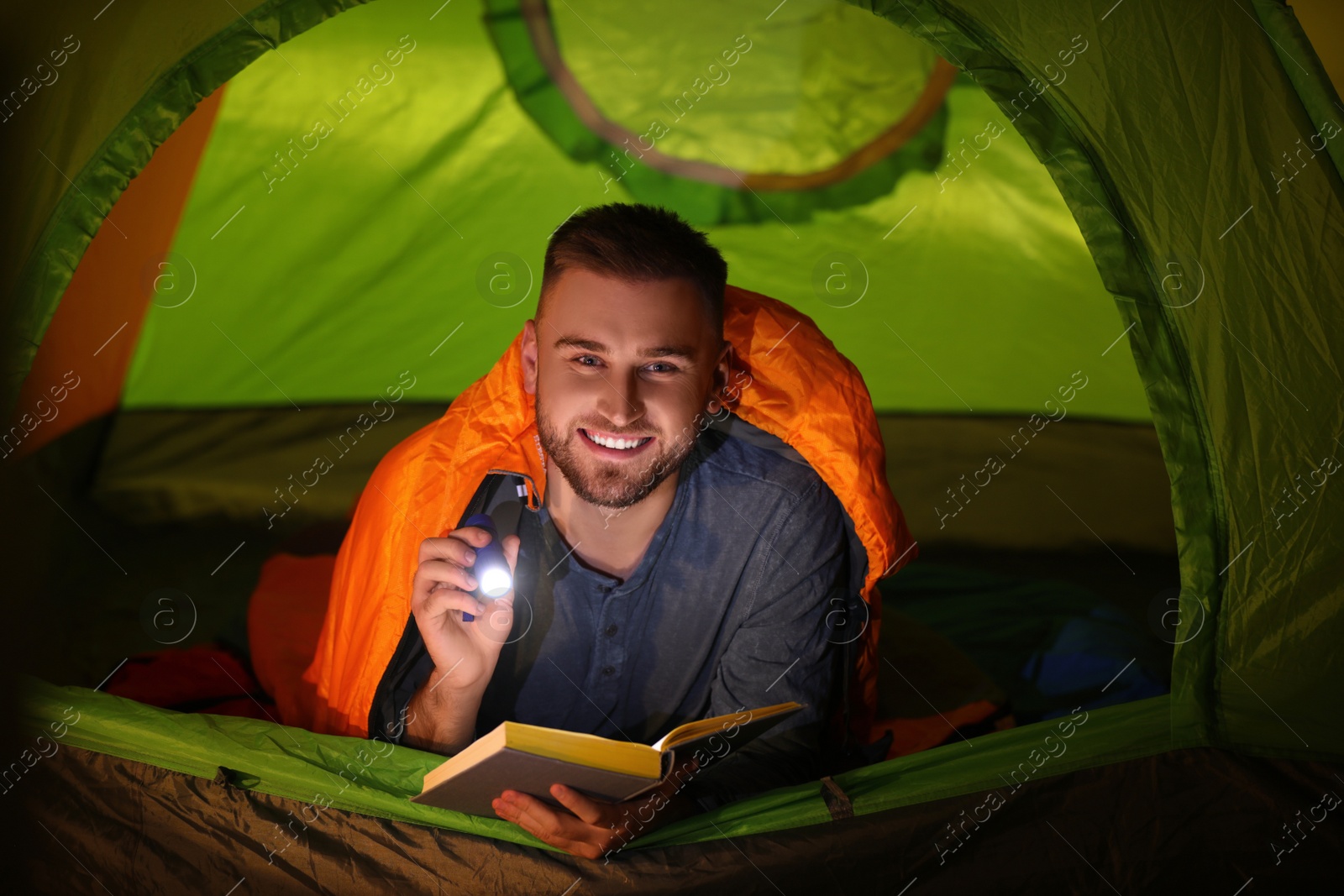 Photo of Young man with flashlight reading book in tent