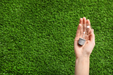 Woman holding incandescent light bulb over green grass, top view. Space for text