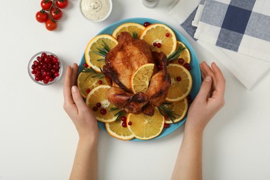 Woman holding plate with baked chicken and orange slices at white table, top view