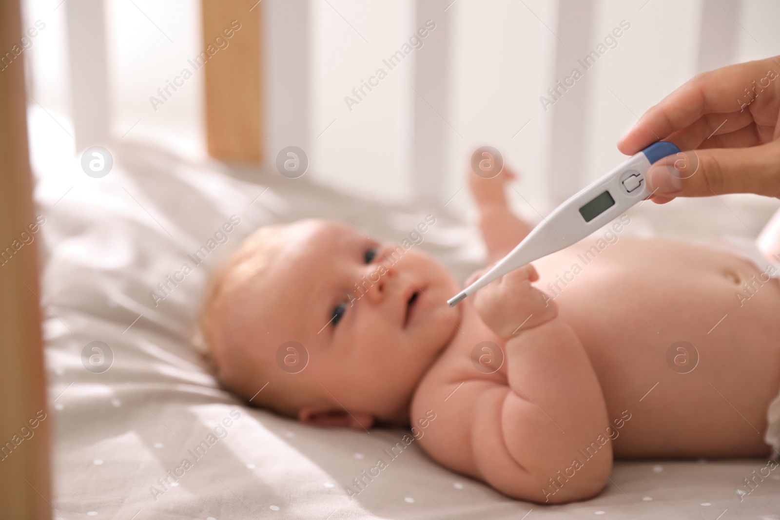 Photo of Cute baby lying in crib, focus on woman holding digital thermometer. Health care