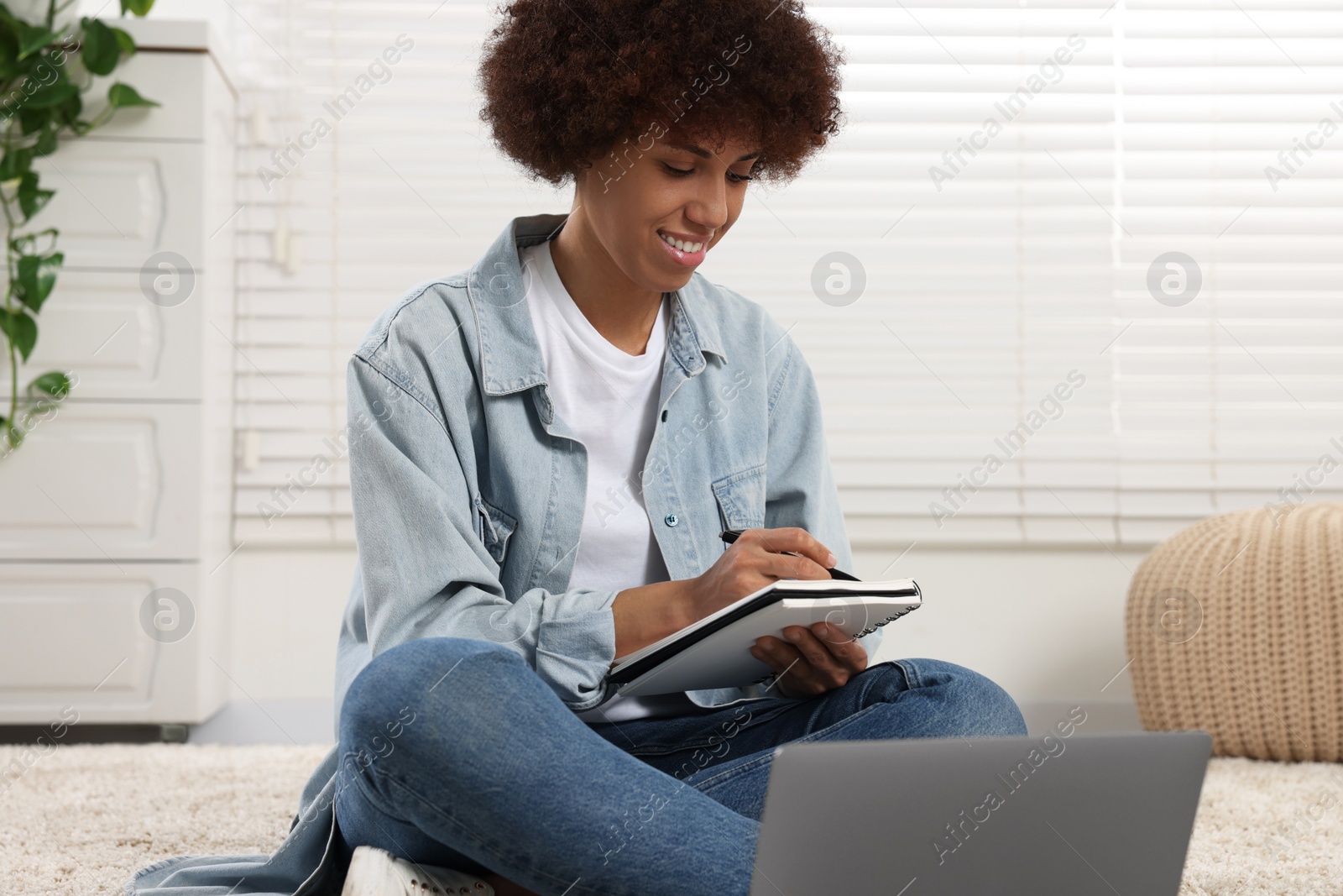 Photo of Beautiful young woman using laptop and writing in notebook in room