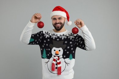 Photo of Happy young man in Christmas sweater and Santa hat holding festive baubles on grey background