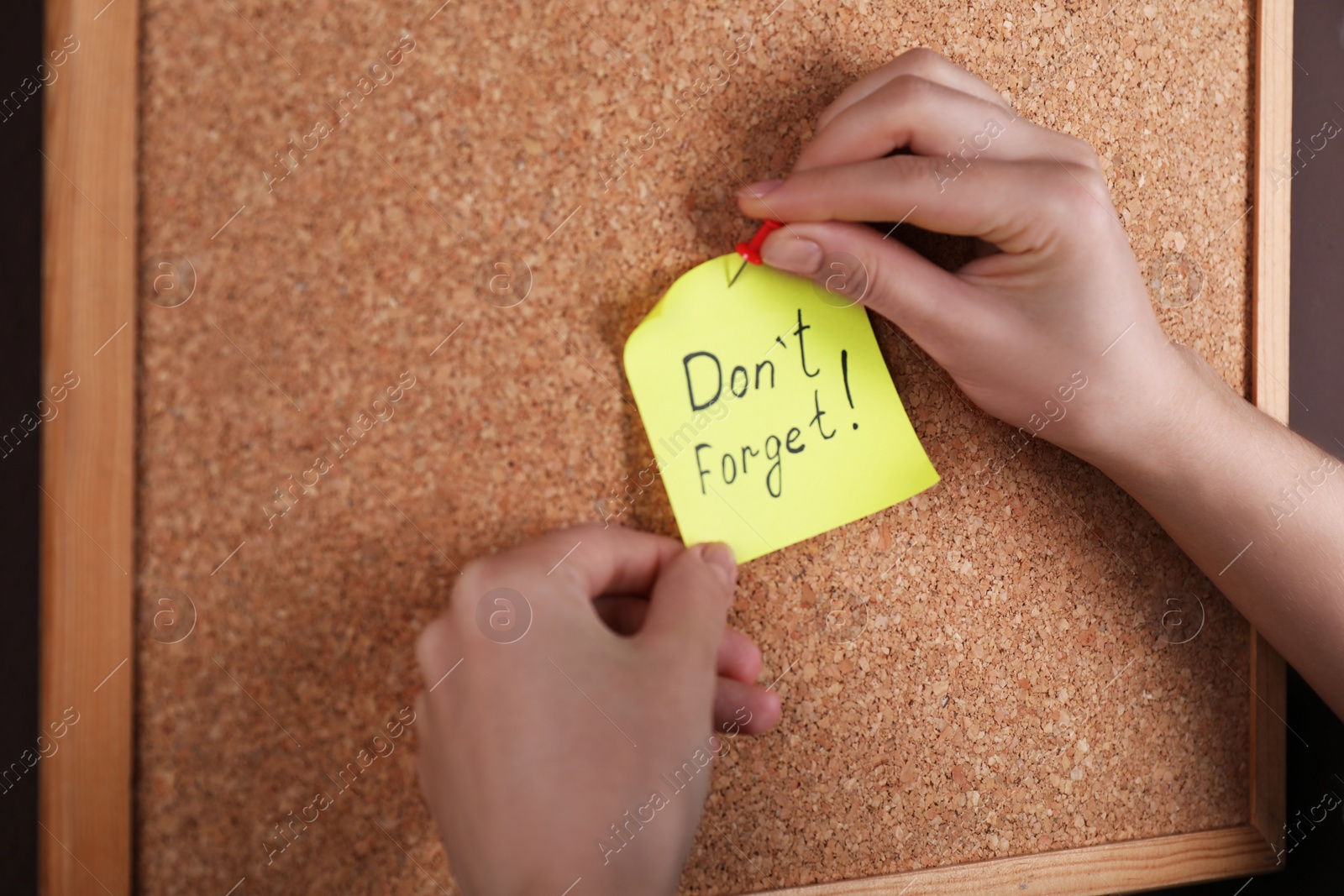 Photo of Woman pinning paper note with phrase Don't Forget to cork board, closeup