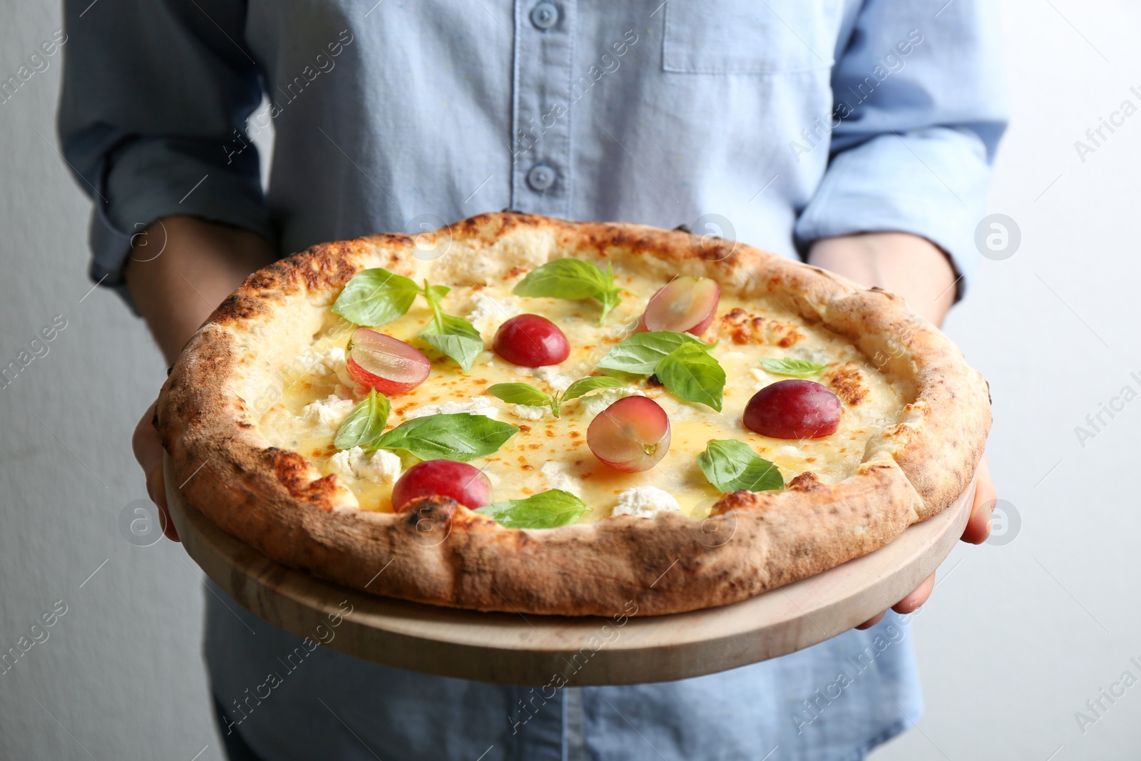 Photo of Woman holding delicious cheese pizza with grapes and basil on light background, closeup