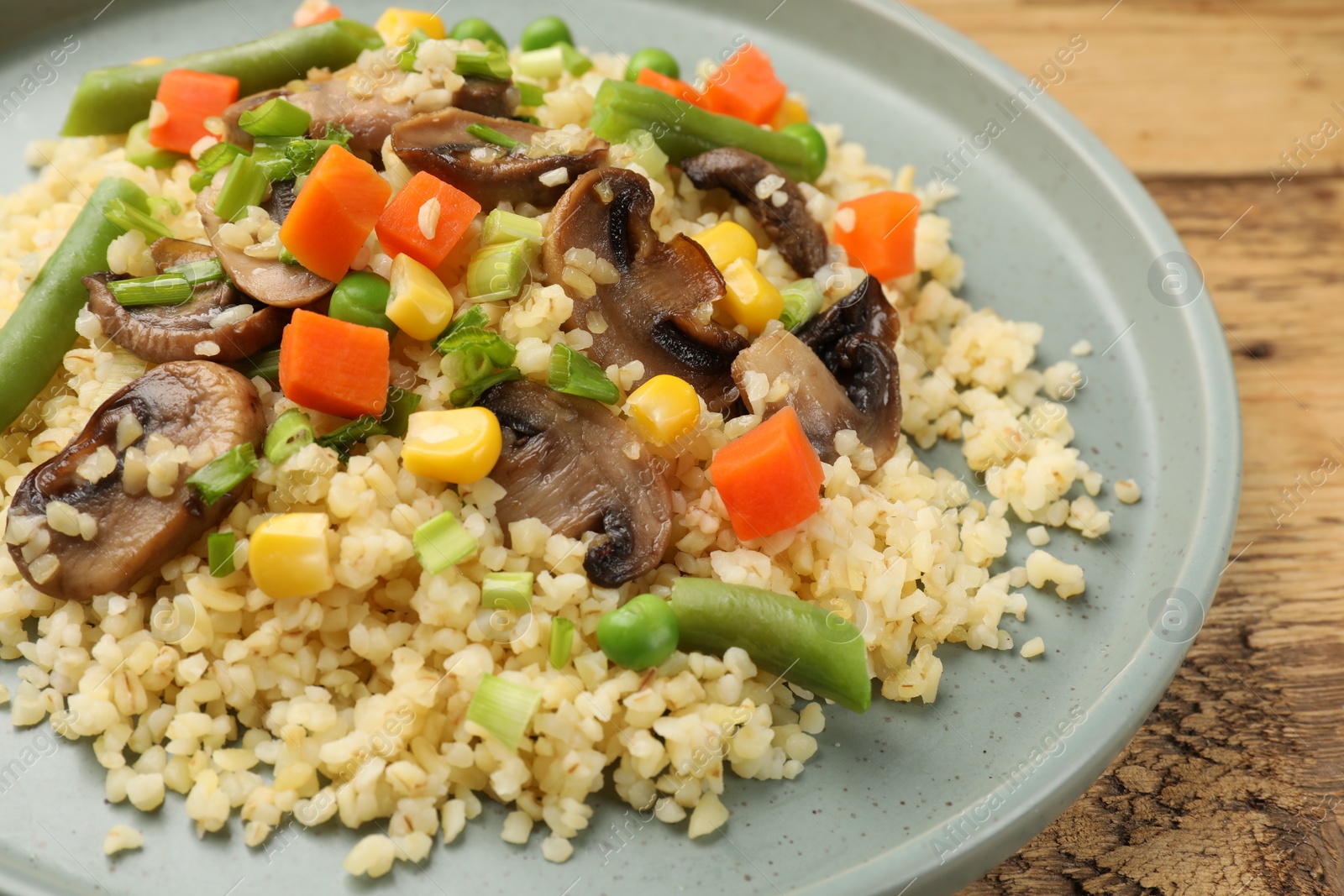 Photo of Delicious bulgur with vegetables and mushrooms on wooden table, closeup