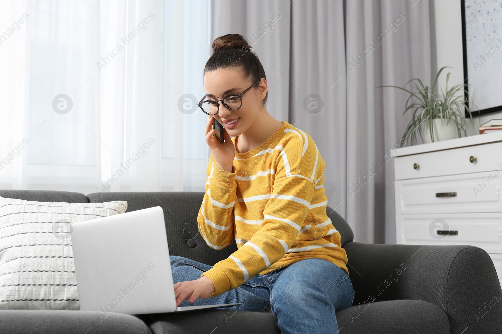 Photo of Home workplace. Happy woman talking on smartphone while working on laptop in room