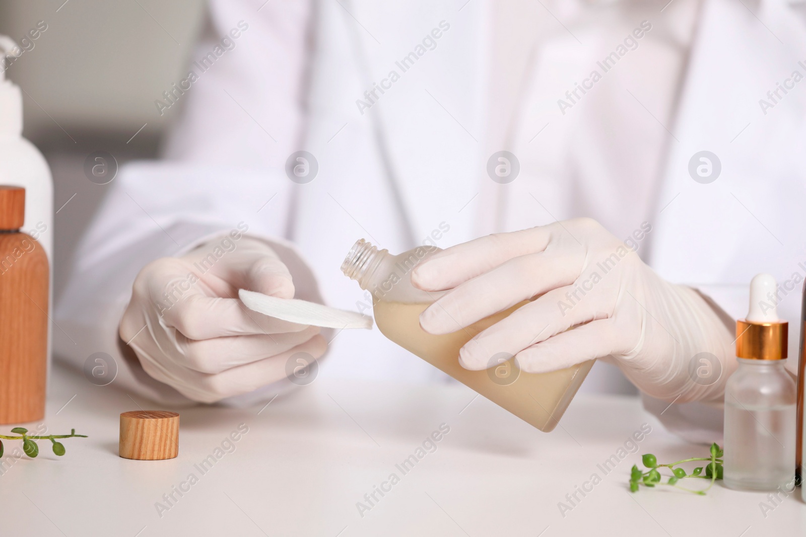 Photo of Dermatologist with bottle testing cosmetic product at white table indoors, selective focus