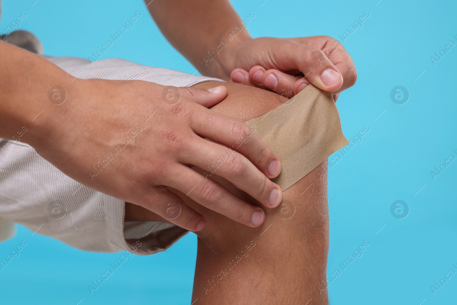Photo of Man putting sticking plaster onto knee on light blue background, closeup