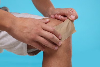 Man putting sticking plaster onto knee on light blue background, closeup