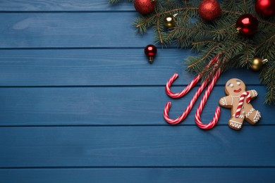 Tasty candy canes, gingerbread cookie, Christmas balls and fir branches on blue wooden table, flat lay. Space for text