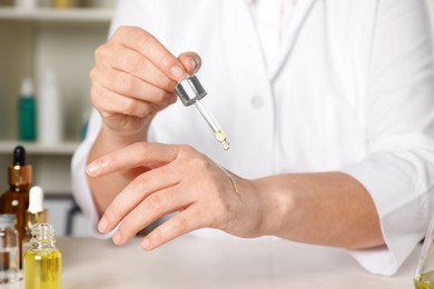 Photo of Dermatologist testing cosmetic oil at white table indoors, closeup