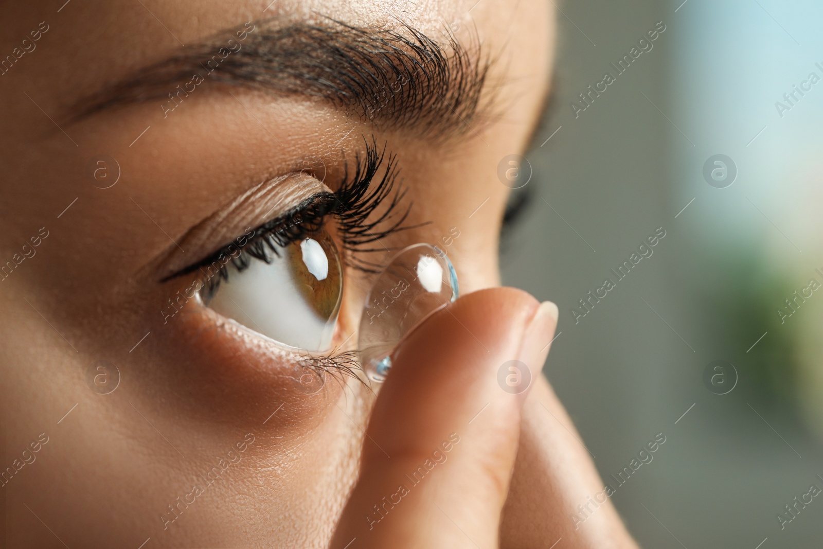 Photo of Young woman putting contact lens in her eye on blurred background, closeup