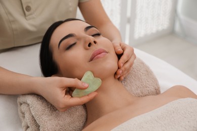 Photo of Young woman receiving facial massage with gua sha tool in beauty salon