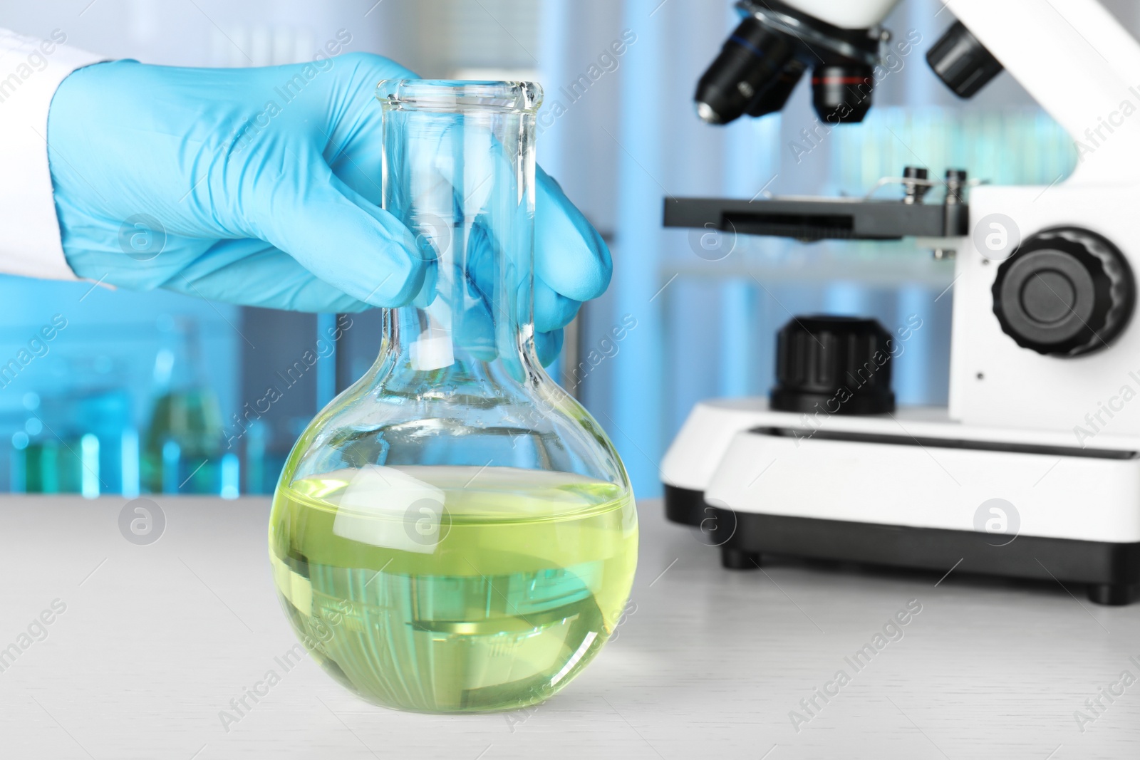 Photo of Scientist holding Florence flask with liquid at white table, closeup