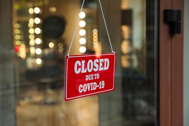 Photo of Red sign with words Closed Due To Covid-19 hanging on glass door, closeup