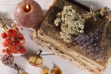 Different dry herbs, flowers, old book and burning candle on white wooden table, flat lay